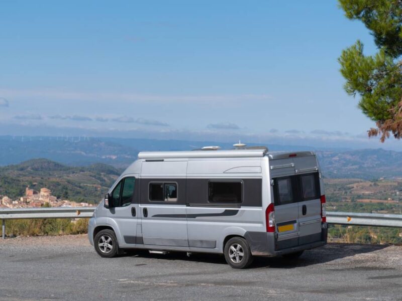 A silver conversion van is parked near a scenic overlook in Spain. In the distance, a town, mountains, and fields are visible.