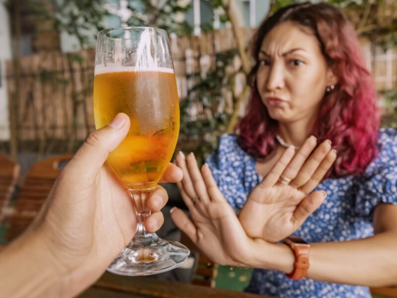 A hand holds a glass of beer out to a woman who has her arms crossed in an X infront of her.