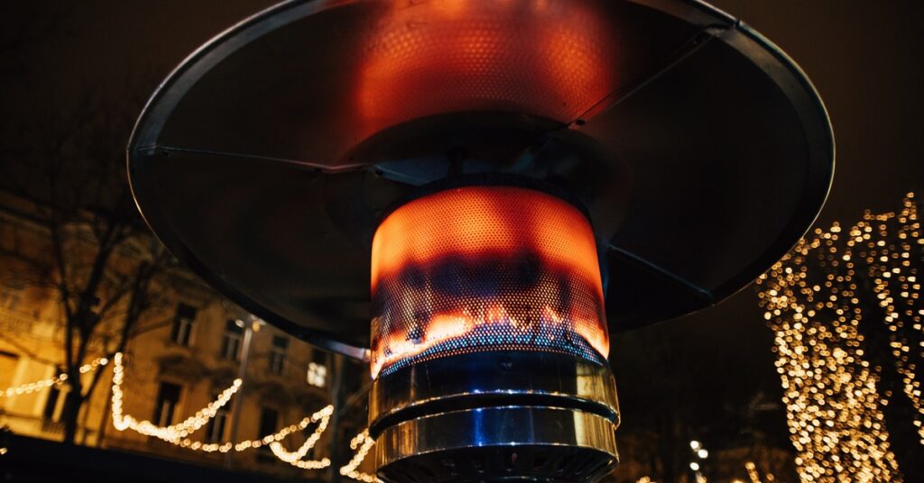A close-up view shows an outdoor heater with a large orange and blue flame. Twinkly lights shine in the background.