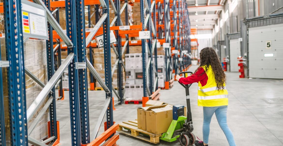 A woman wearing a hi-visibility yellow vest pushes a pallet jack into an aisle between two orange and blue pallet racks.
