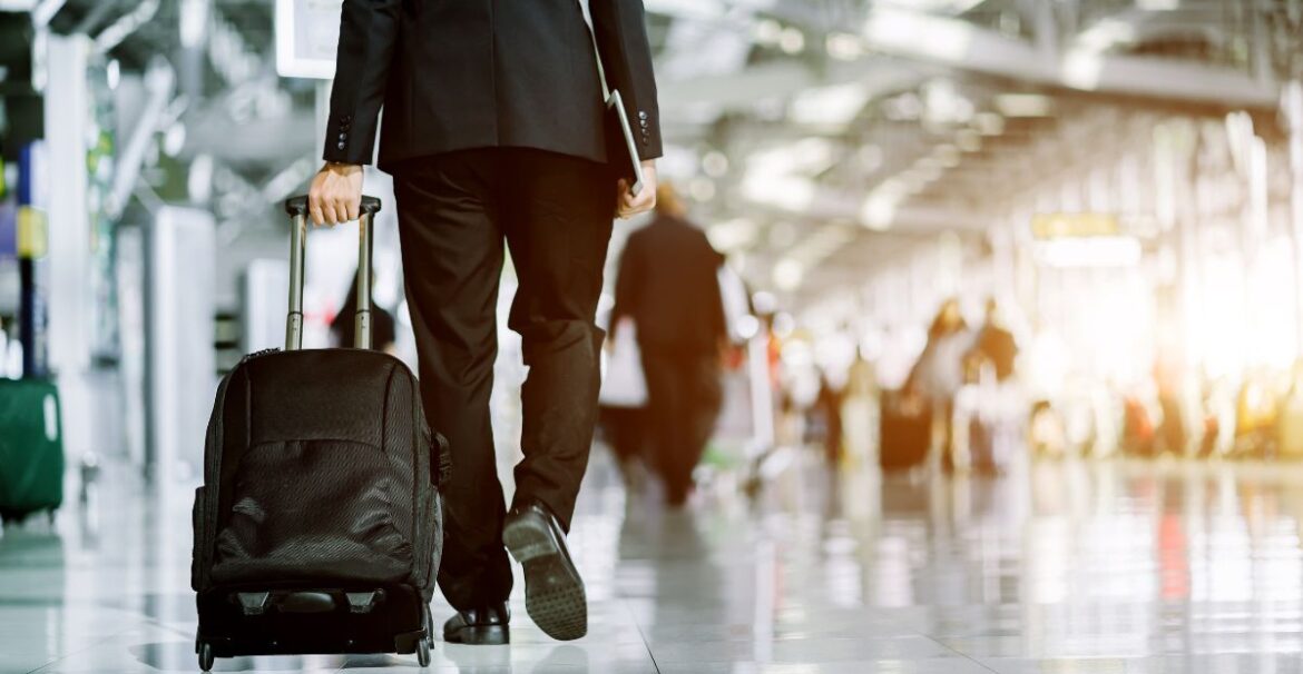 Rear view of a business person walking through an airport with a roller suitcase behind him. Other travelers are around him.