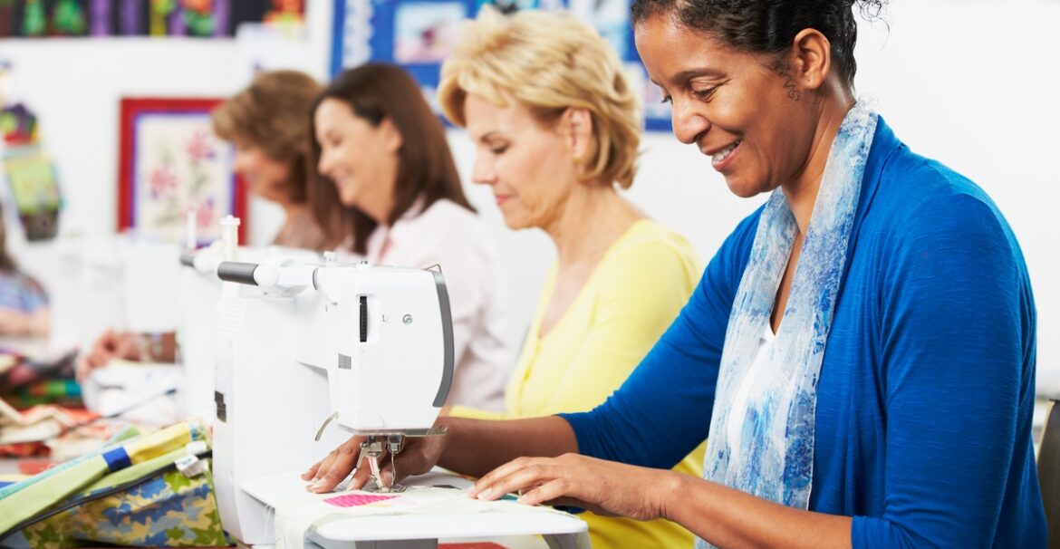 Four women sit next to each other at a table sewing. Each woman is looking down at their sewing machine.