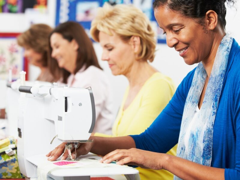 Four women sit next to each other at a table sewing. Each woman is looking down at their sewing machine.