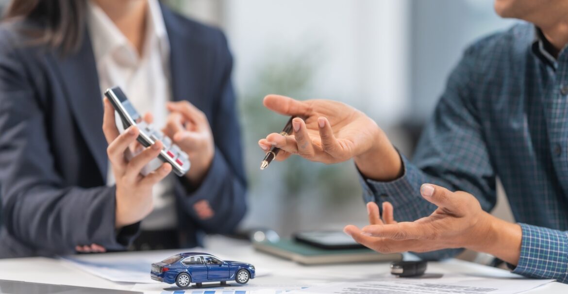 Two people sit together at a desk across from one another. One holds a calculator over a toy car and stacks of paper.