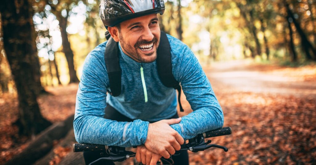 A man wearing a helmet and blue sweatshirt smiles while leaning on a mountain bike along a forest trail.