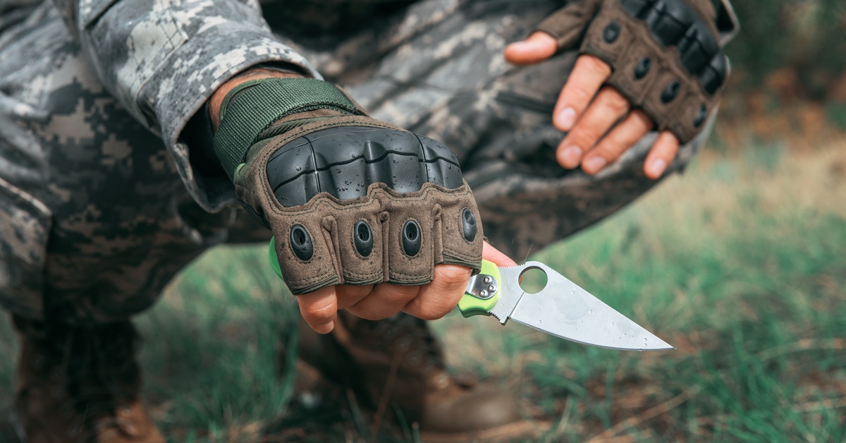 A man is crouching and holding a small knife in the forest. He wears military gear, including gloves and boots.