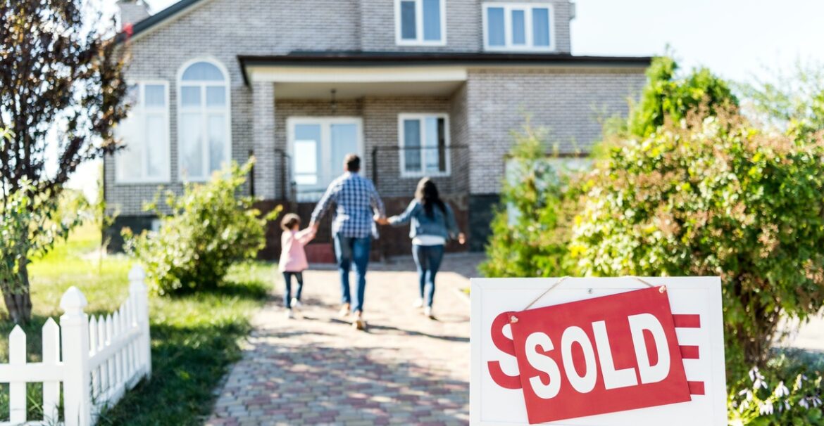 A family holding hands while walking toward a two-story brick house with a bold red sold sign in the yard.