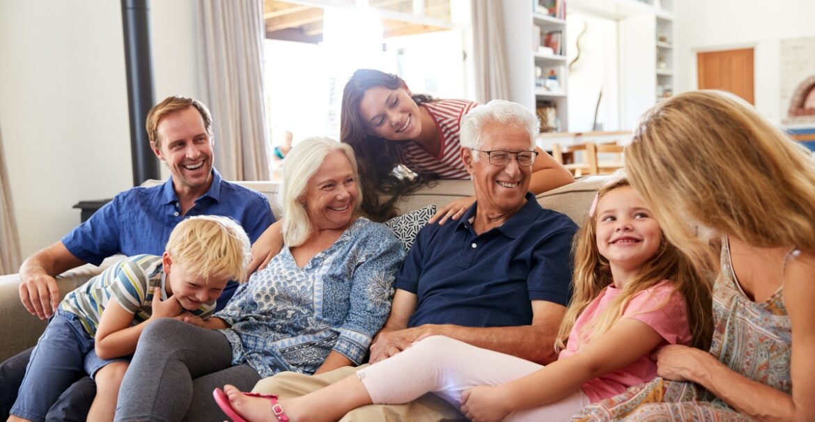 Two young children smile as they sit on a couch with three middle aged adults and two elderly people.