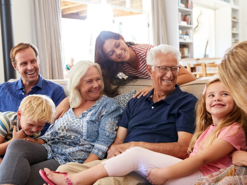 Two young children smile as they sit on a couch with three middle aged adults and two elderly people.