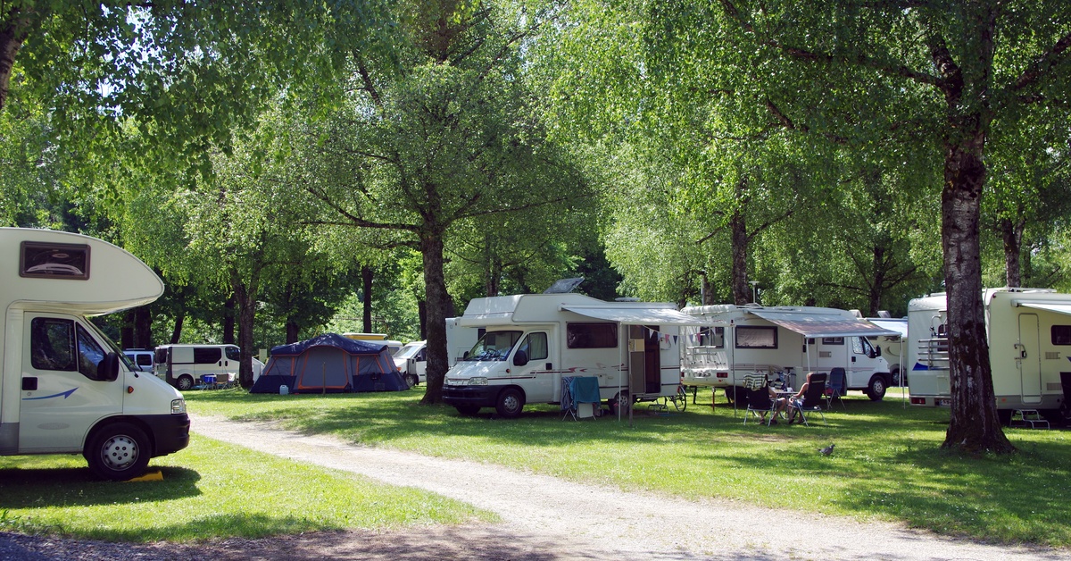A lot of white RVs are parked on green grass surrounded by trees on a campsite with a blue tent set up as well.